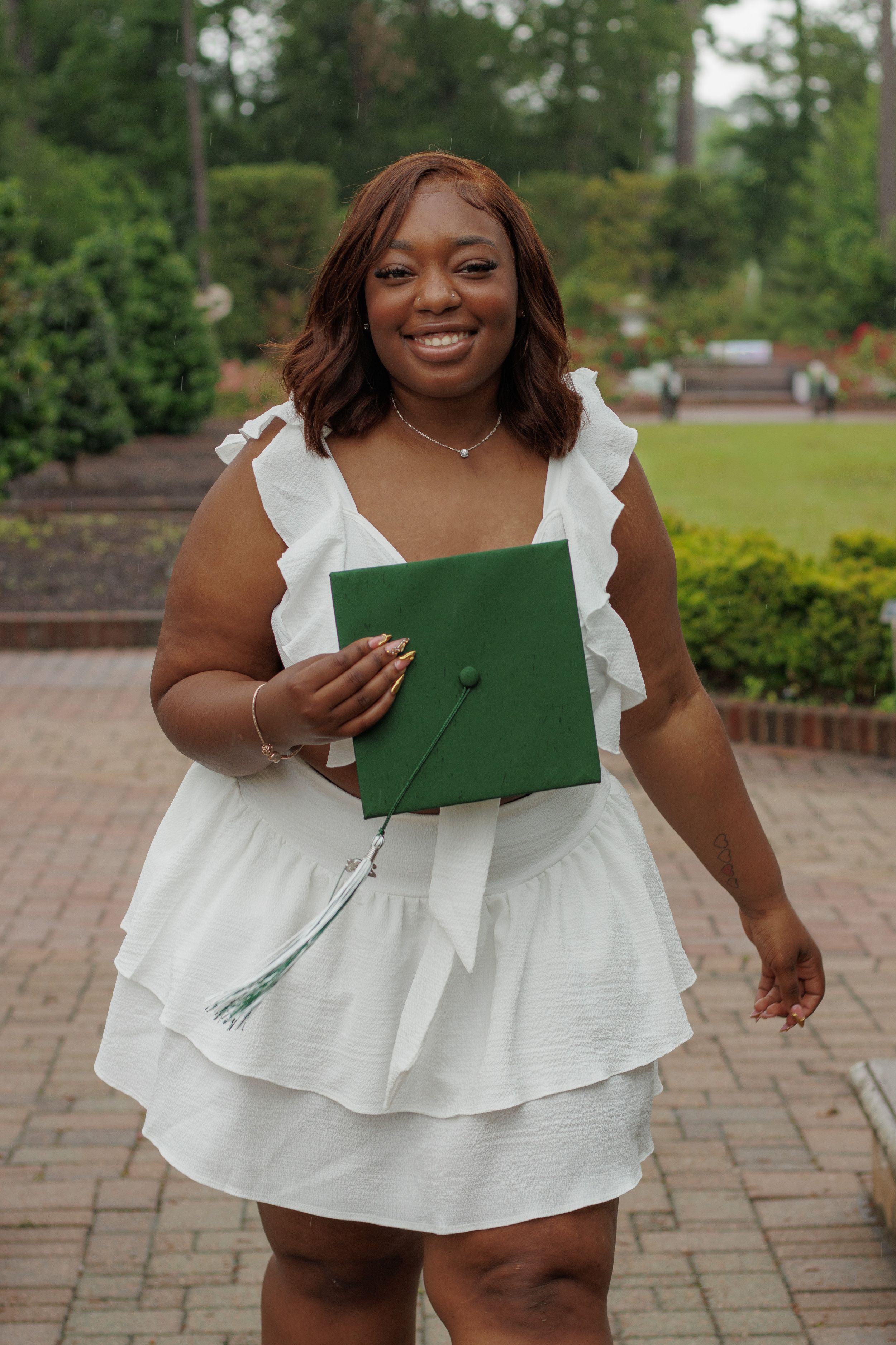 Smiling young woman in white dress holding green graduation cap outdoors on brick path with lush greenery.