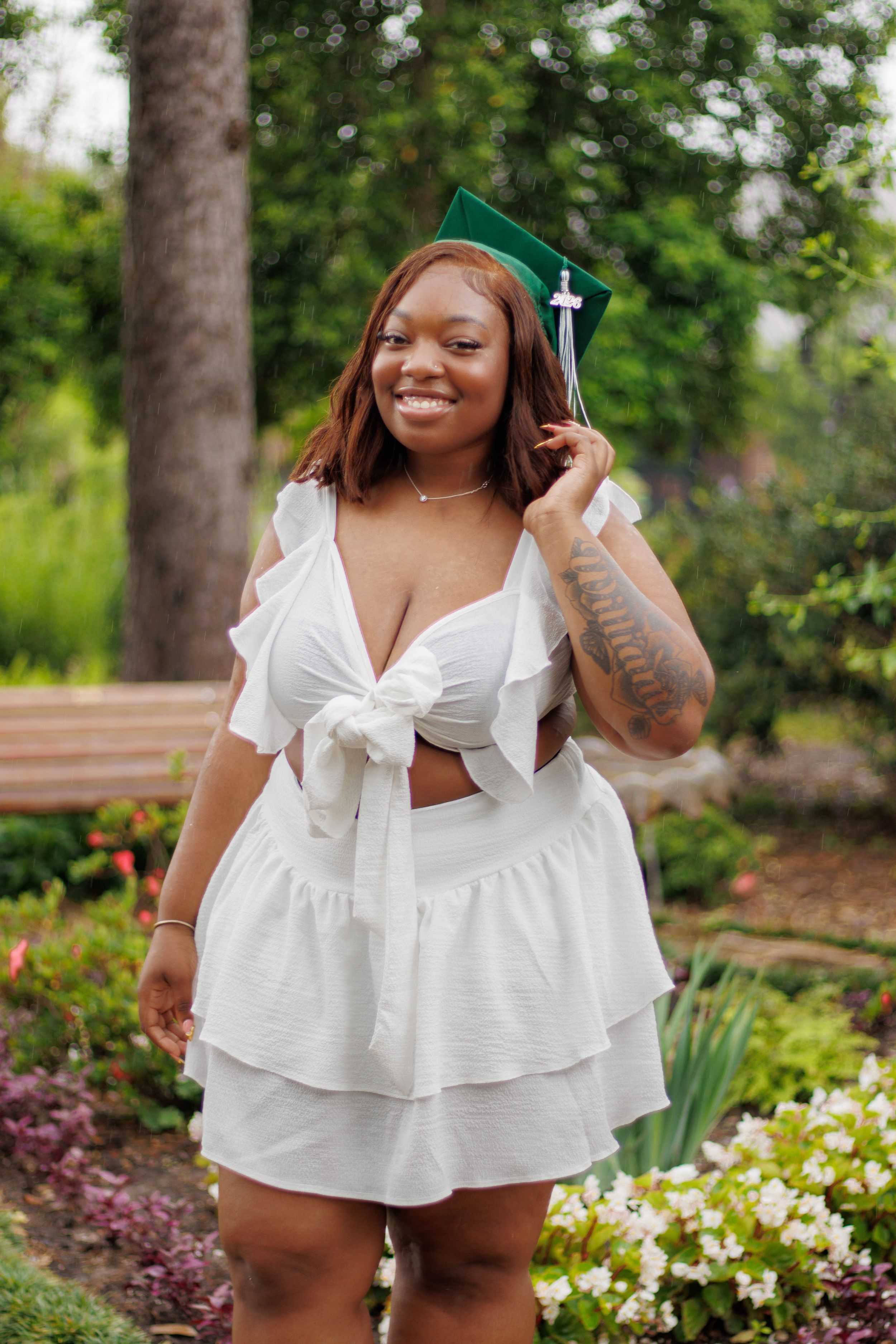 Smiling graduate in white ruffled outfit holding green cap in a garden setting.