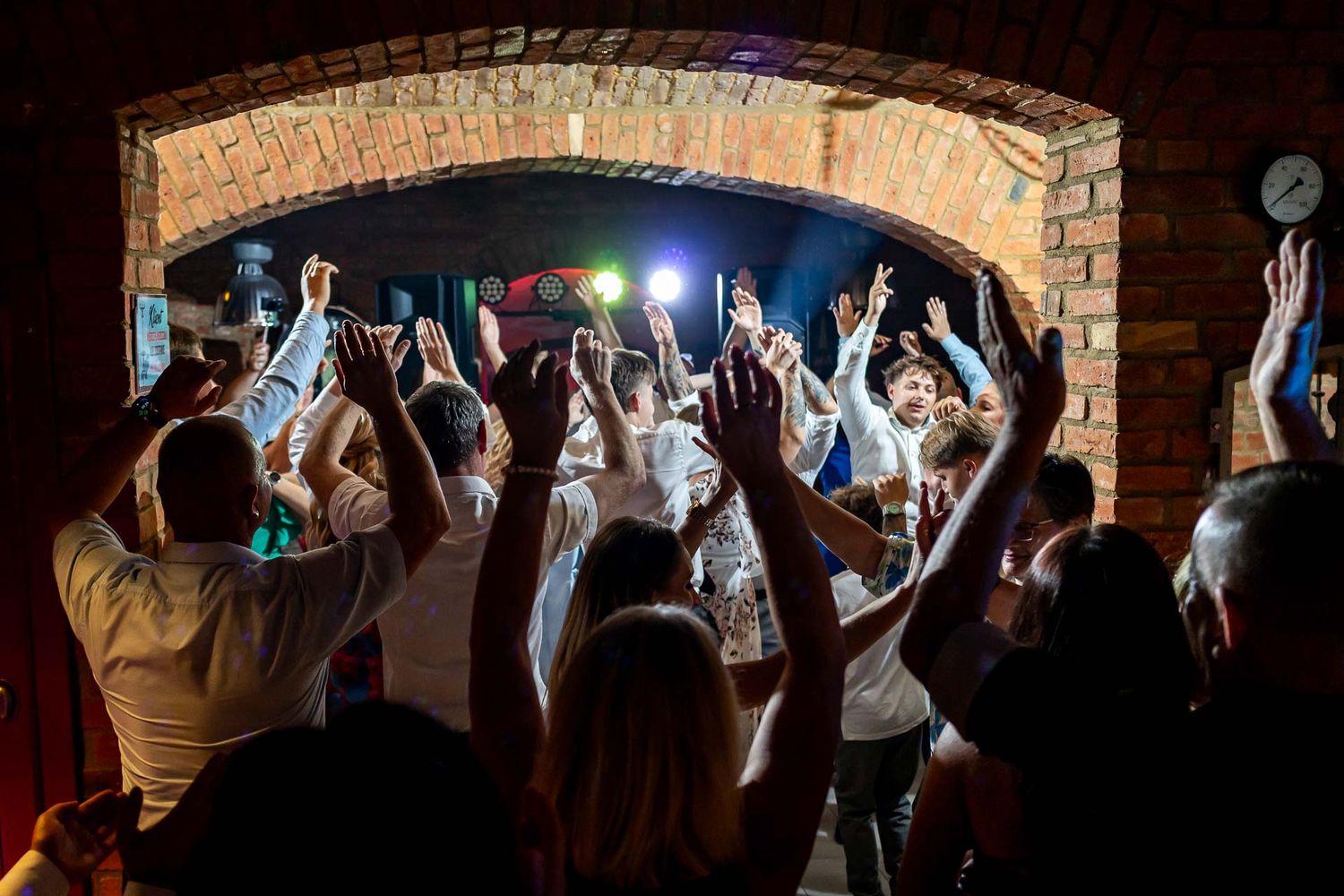 Wedding guests with raised hands dancing in a dimly lit brick-walled venue.