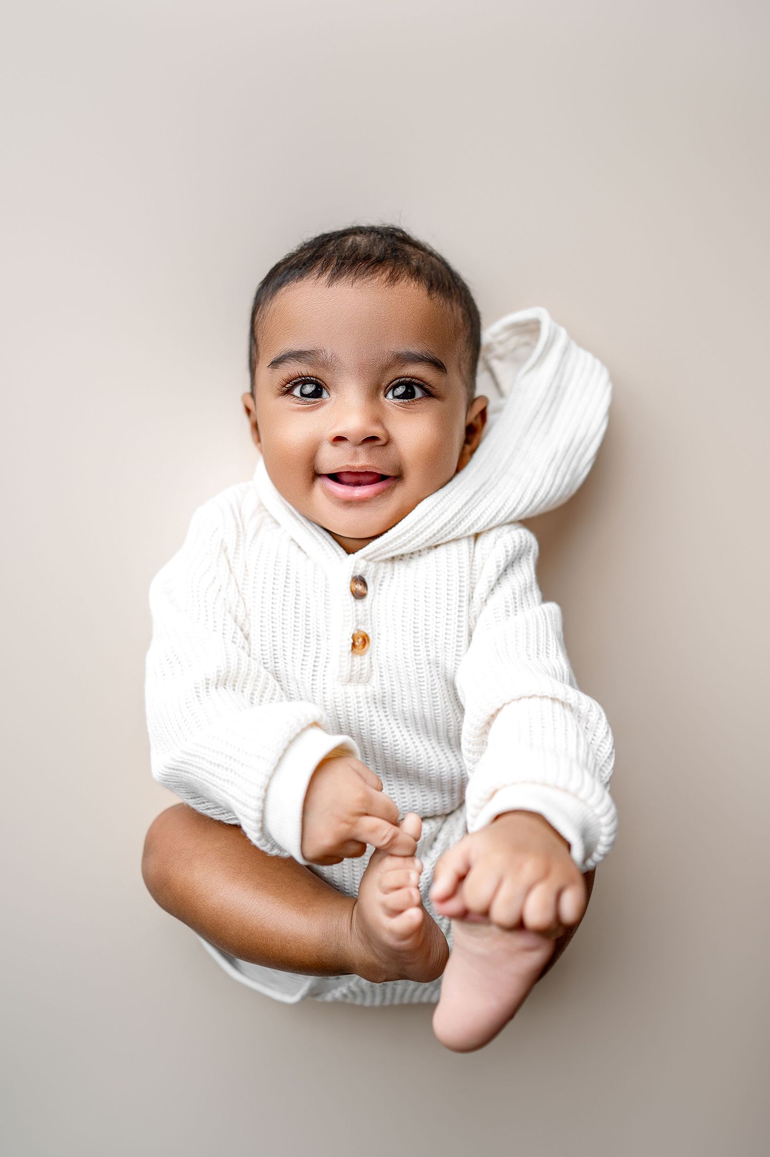 Smiling Black baby in white ribbed hoodie romper lying down, holding feet, looking up with bright eyes.