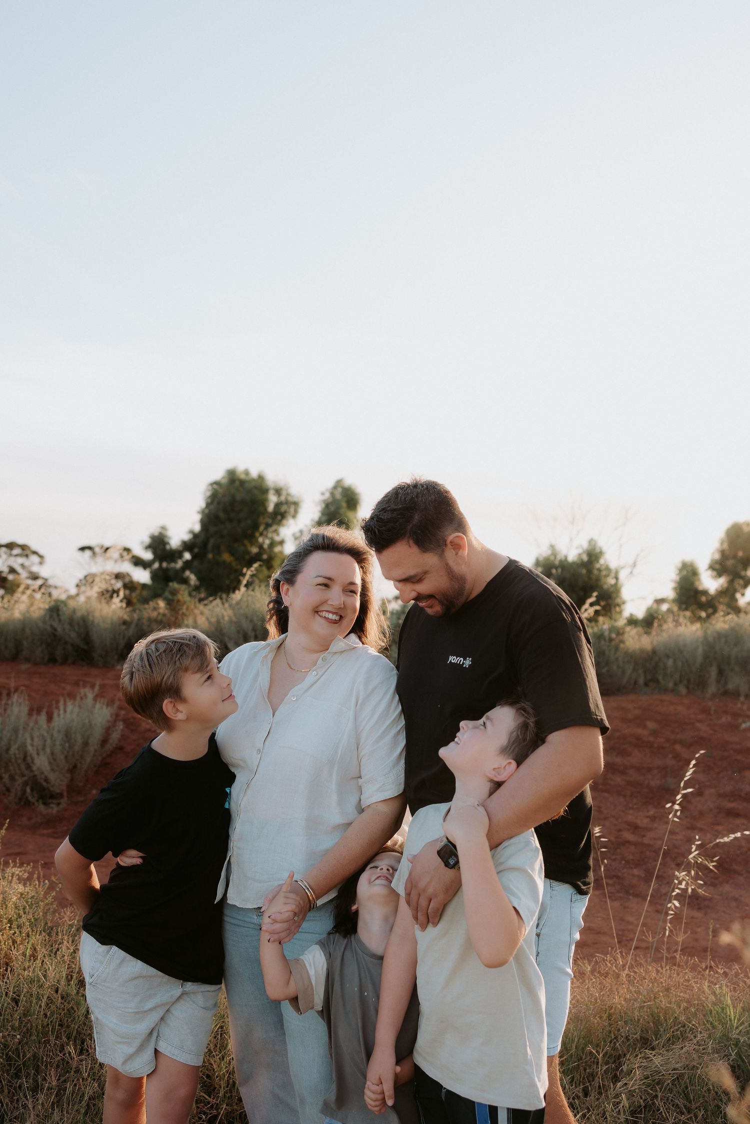 Family of four laughing together in a sunlit field with red earth and green shrubs in the background.