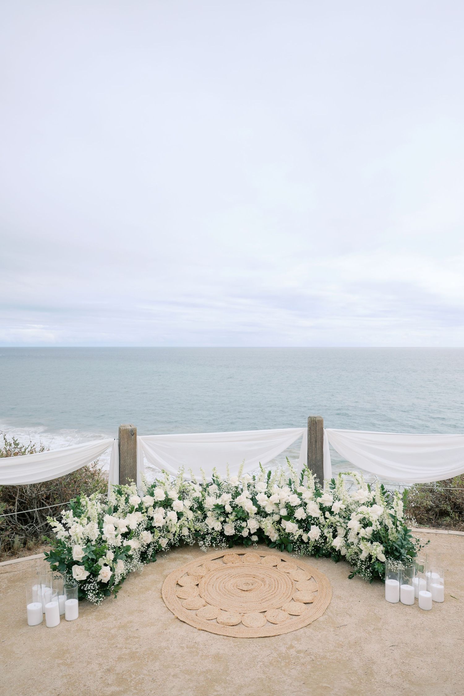 Elegant outdoor wedding altar with white floral arch, draped fabric, and candles overlooking a serene ocean backdrop.