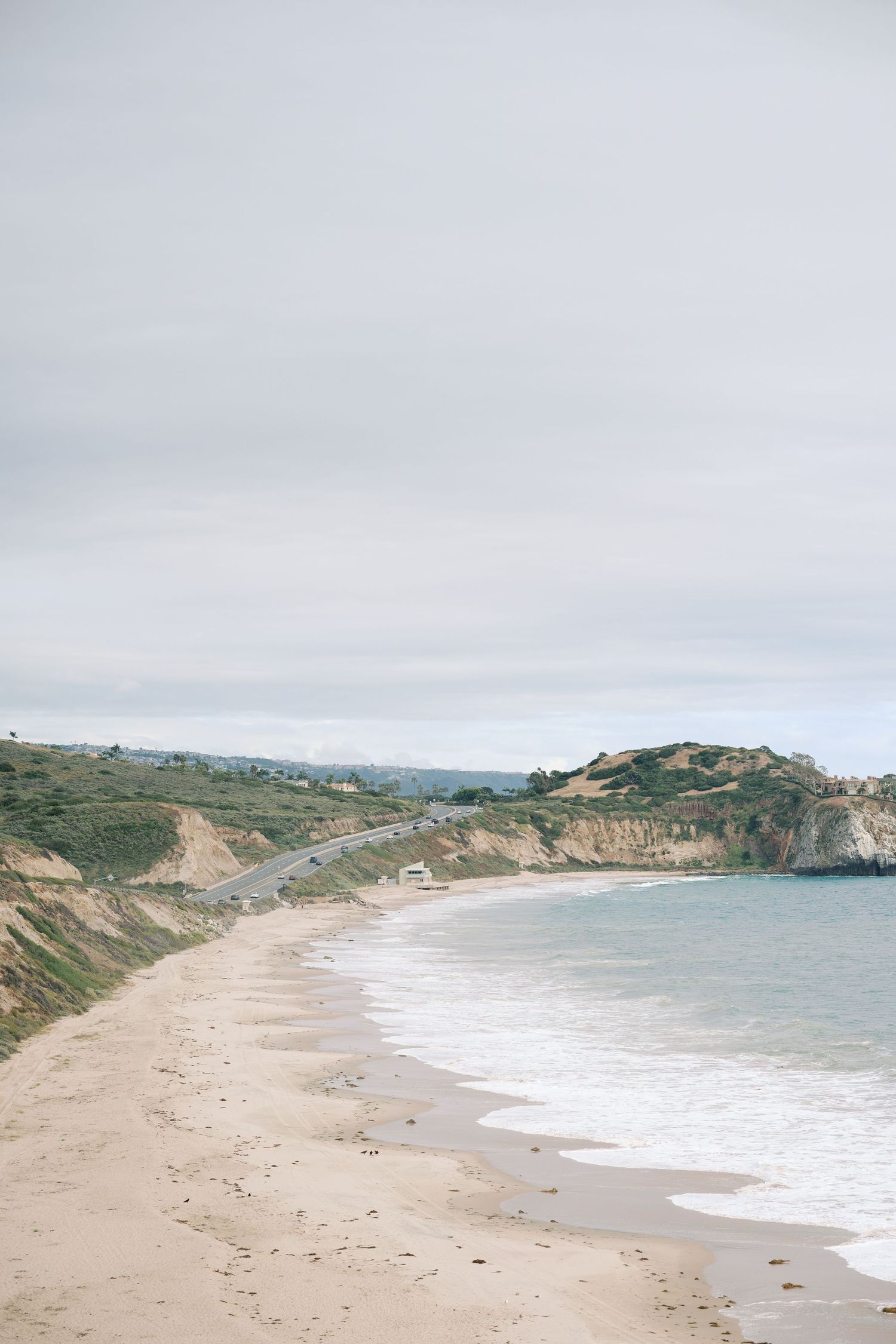 Sandy beach curves along rugged coastal cliffs under overcast skies, with gentle waves and a scenic highway in the background.