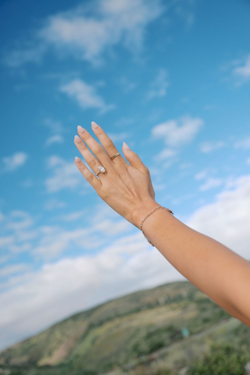 A woman's hand adorned with a gemstone ring and delicate bracelet raised against a blue sky with green hills.