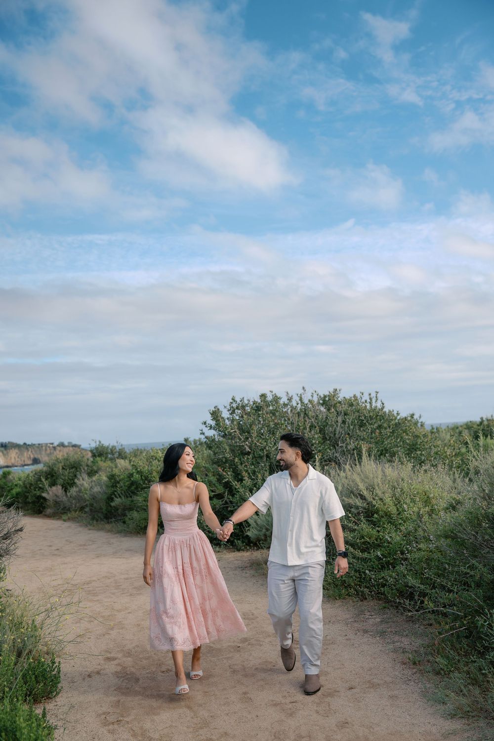 Couple holding hands walking on a scenic coastal path, woman in pink dress, man in white shirt, blue sky background.