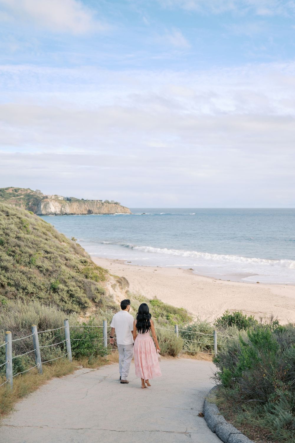 Couple holding hands walking down a coastal path toward a sandy beach with ocean views and cliffs.