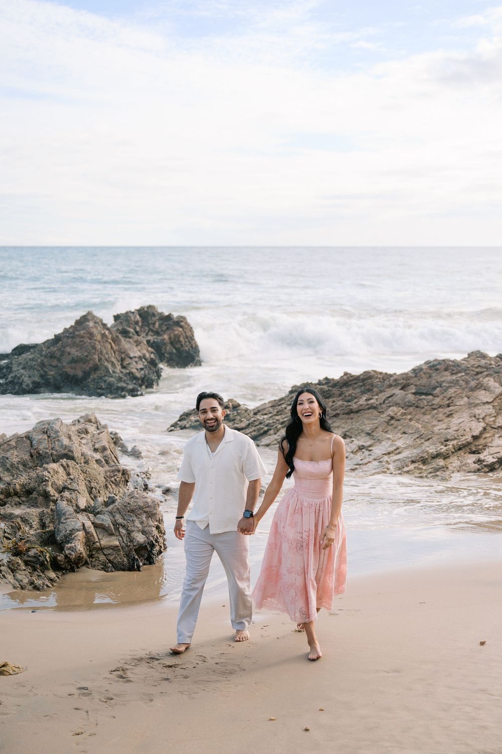 A happy couple holds hands walking barefoot on a sandy beach with rocky shores and ocean waves behind them.