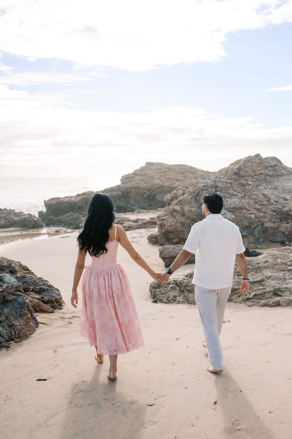 Couple holding hands walking barefoot on sandy beach toward rocky shore, woman in pink dress, man in white shirt and pants.