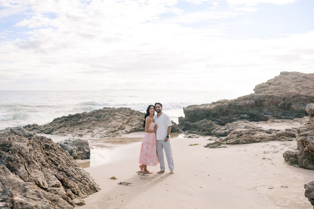 Couple posing together on a rocky beach with ocean waves and cloudy sky, woman in pink dress, man in white shirt.