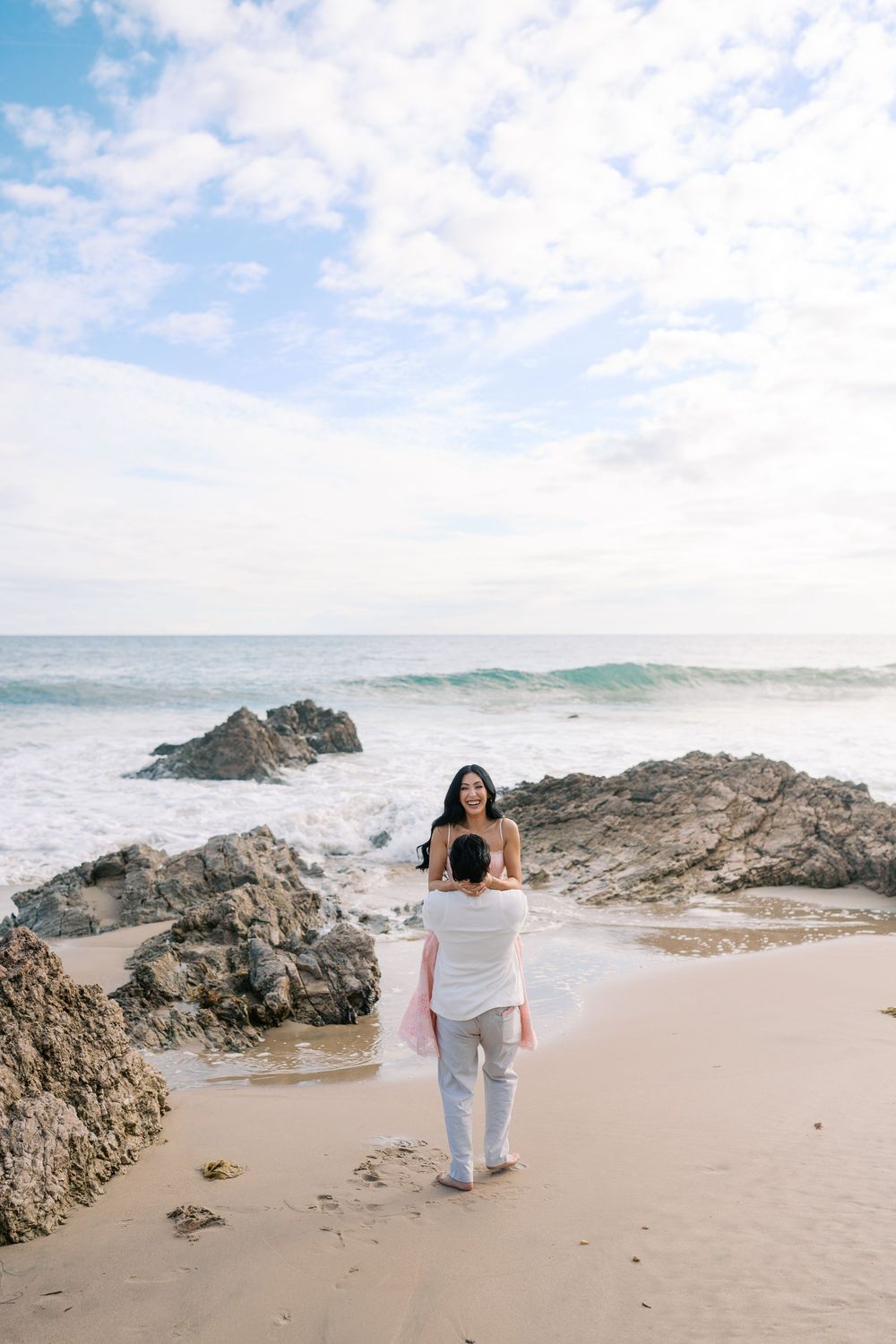 Couple on a rocky beach, man lifting woman in white dress, ocean waves and cloudy sky in background.