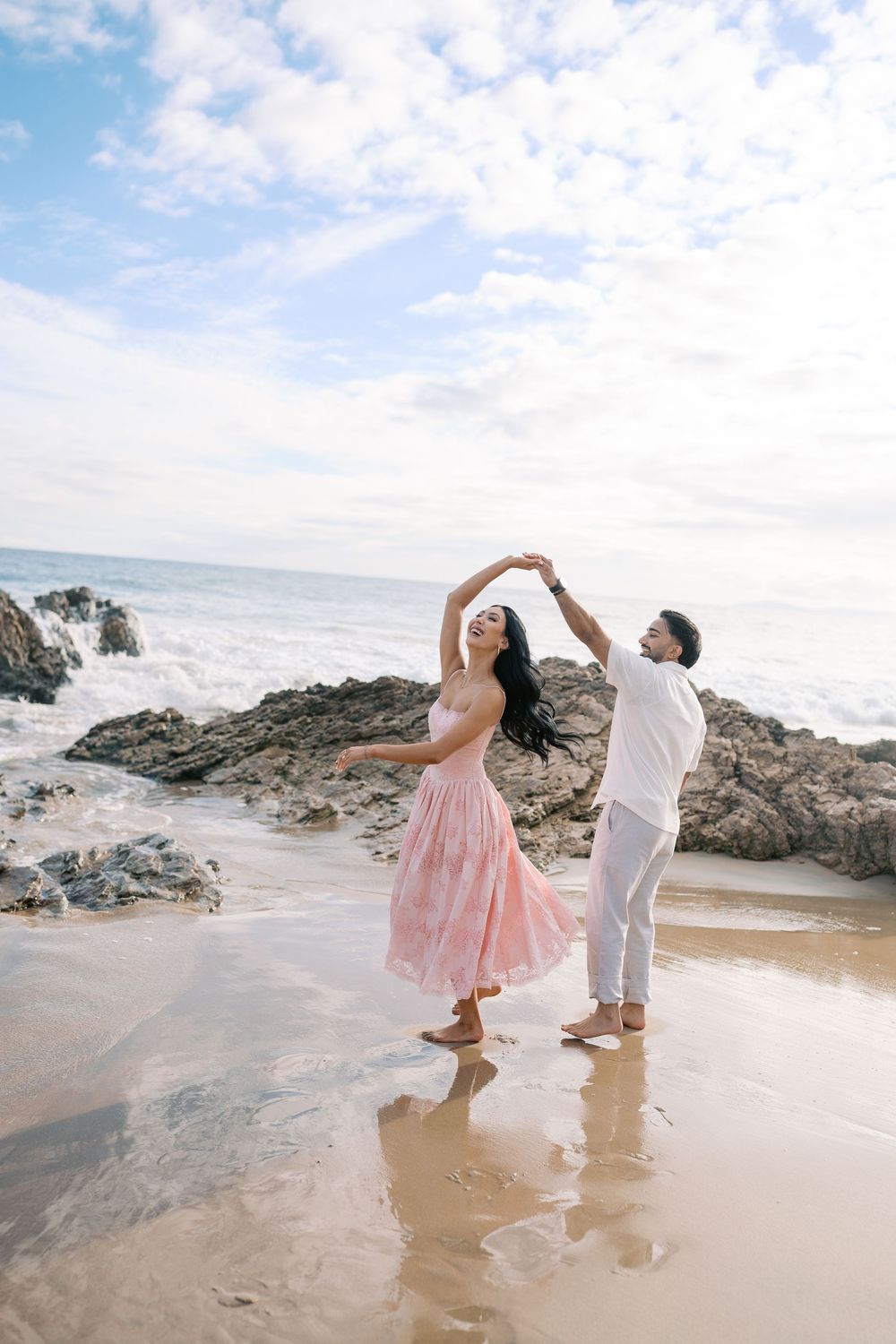 Couple dancing barefoot on a sandy beach, woman in pink dress being twirled, with rocky shore and ocean waves behind them.