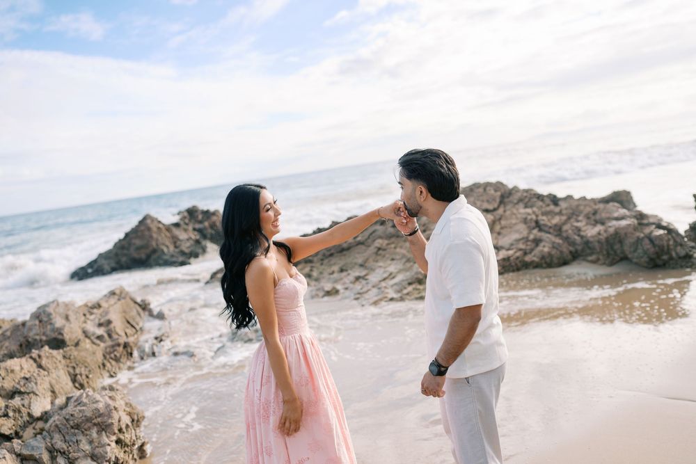 Couple sharing a romantic moment on a rocky beach, woman in pink dress touching man's face with ocean waves behind them.