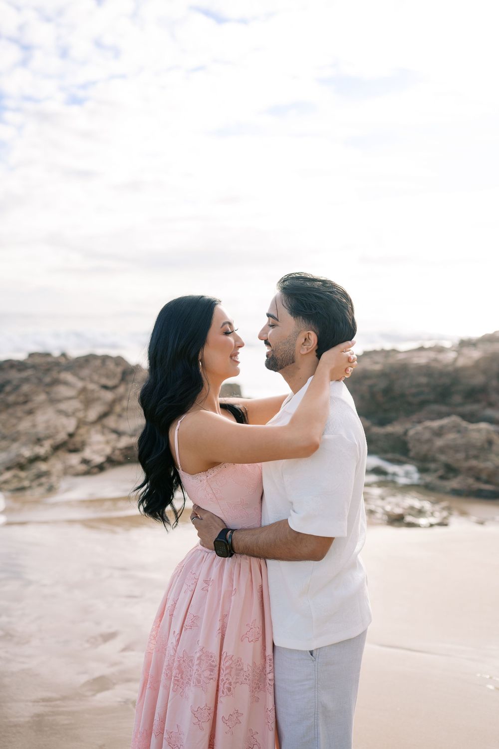 Couple embracing on a beach, woman in pink dress and man in white shirt, sharing a romantic moment by rocky shores.
