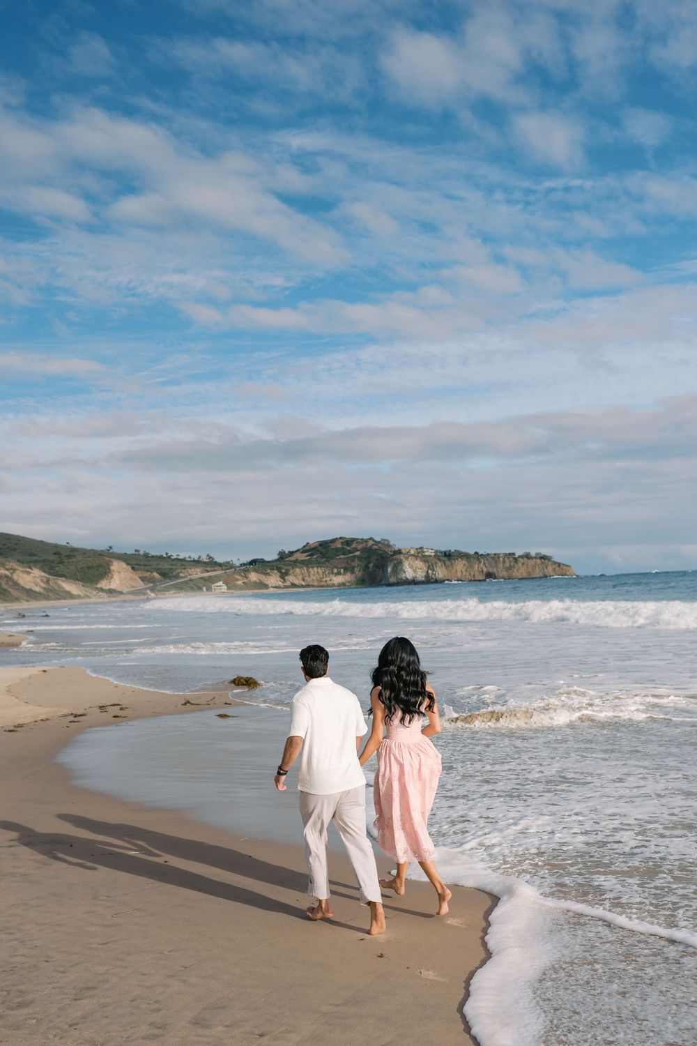 A romantic couple walks hand in hand along a sandy beach shoreline with crashing waves and coastal cliffs in the distance.