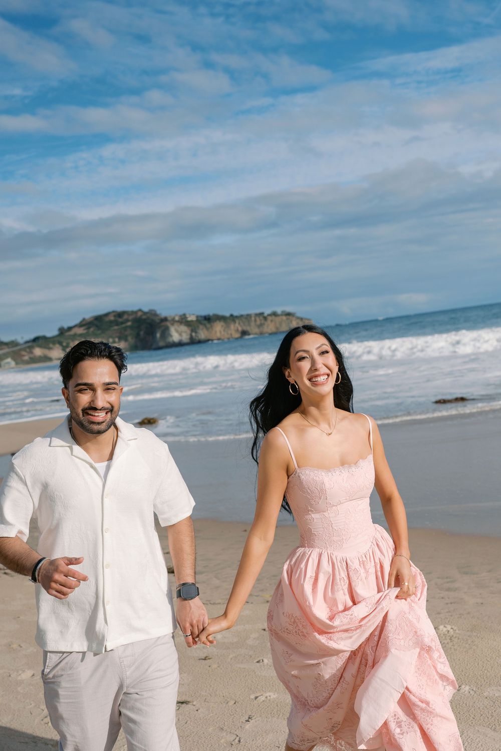 Happy couple holding hands and running on a sunny beach, woman in pink dress and man in white outfit.
