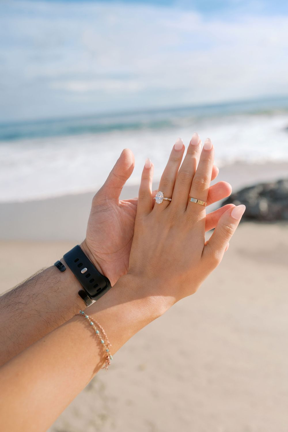 Close-up of engaged couple's hands showing diamond engagement ring on beach with ocean backdrop.