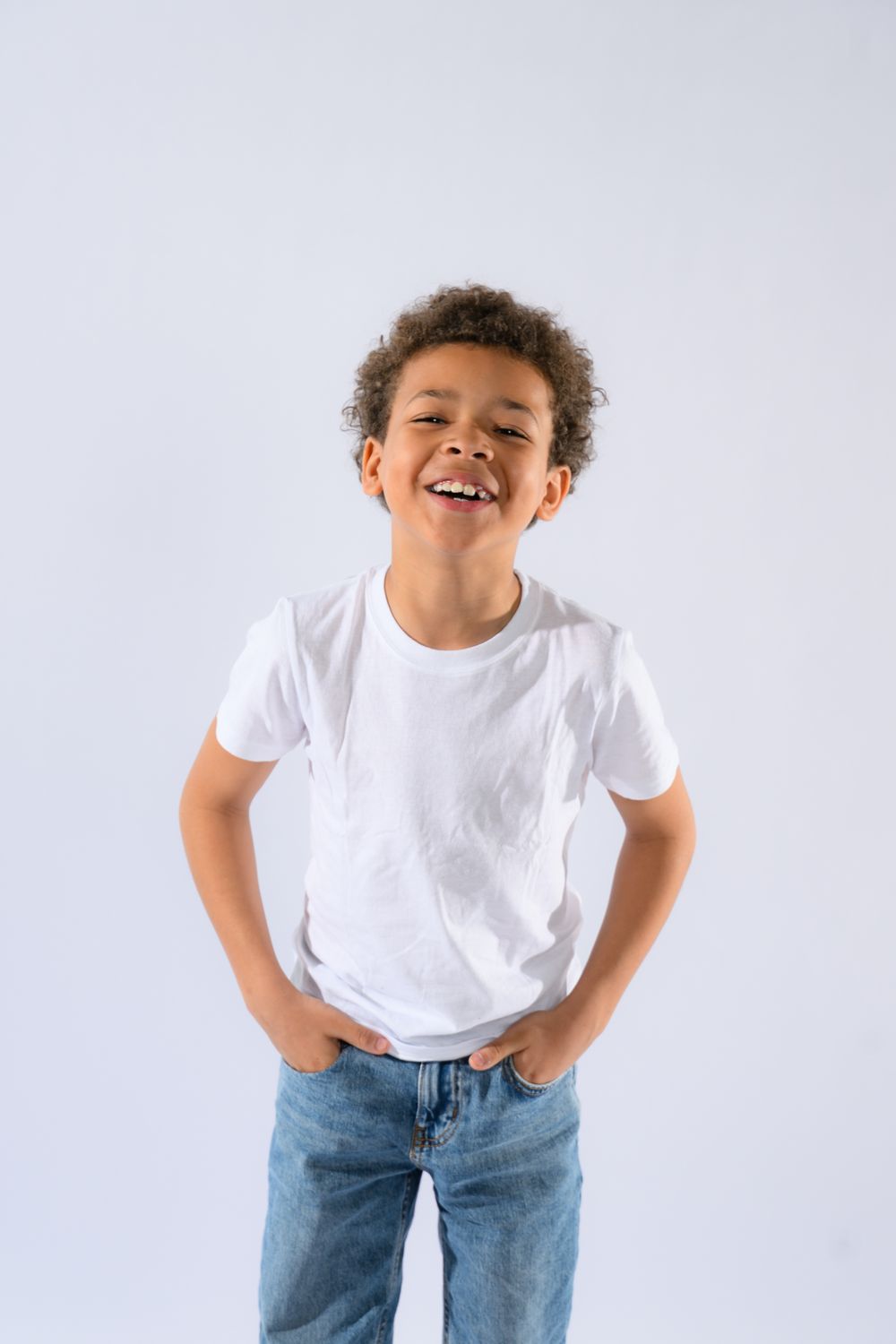 Happy young African American boy in white t-shirt and jeans smiling with hands in pockets on white background.
