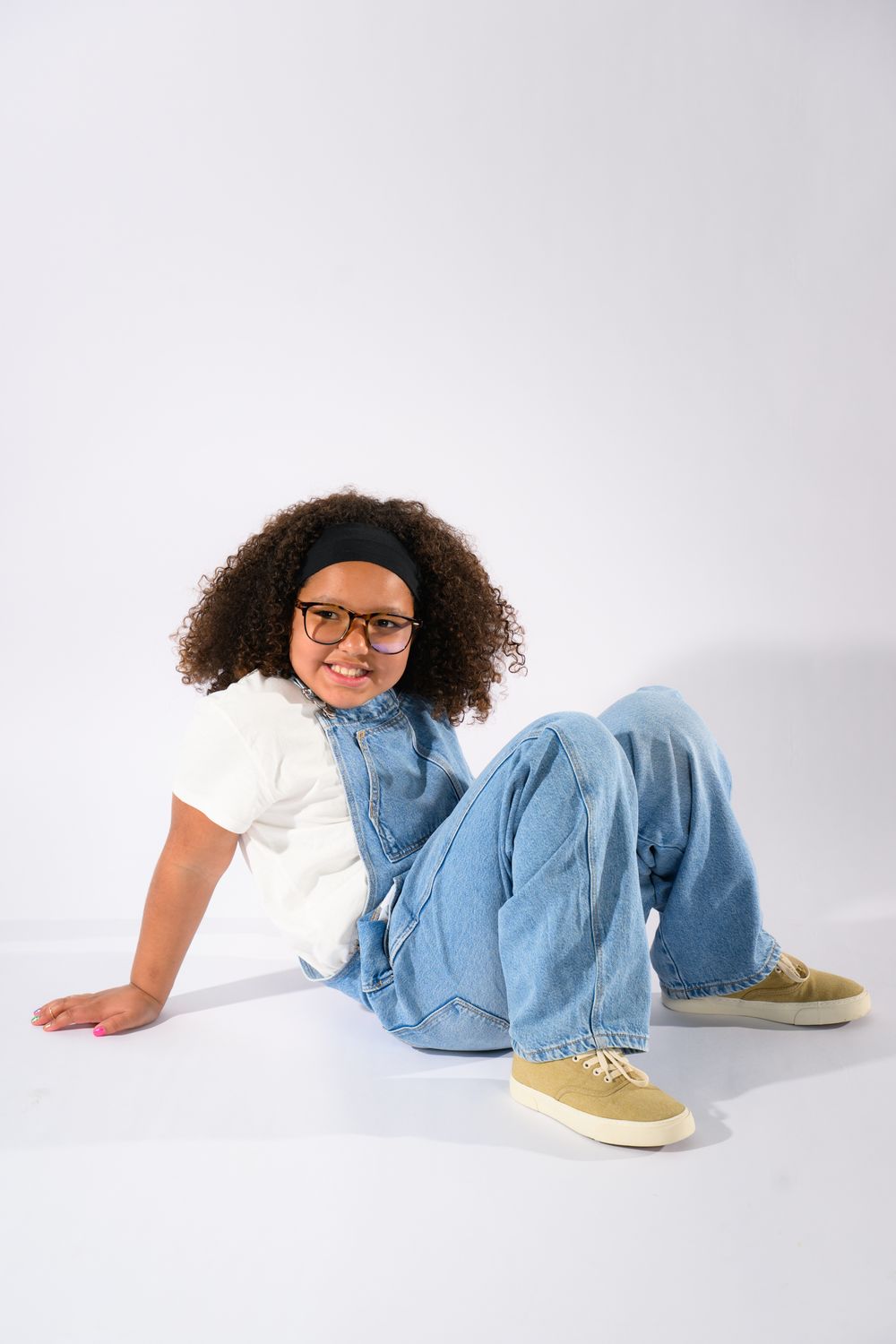 A young girl with curly hair and glasses sits on the floor wearing jeans, a white top, and tan sneakers.