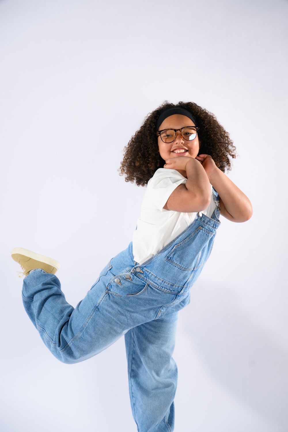 A joyful young girl with curly hair wearing glasses and denim overalls strikes a fun pose on a white background.