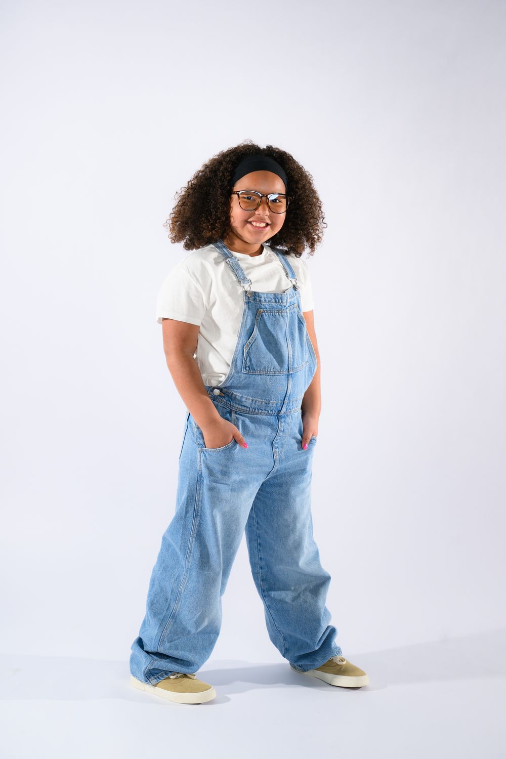 A young girl with curly hair wearing denim overalls, white shirt, and glasses poses confidently on a white background.