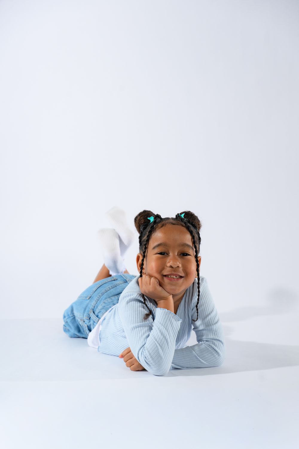 Smiling young Black girl with braided pigtails lying on floor, wearing blue sweater and jeans on white background.