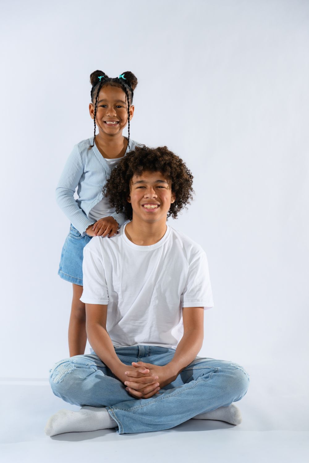 Smiling African American teenage boy sitting cross-legged with young girl standing behind him on white background.