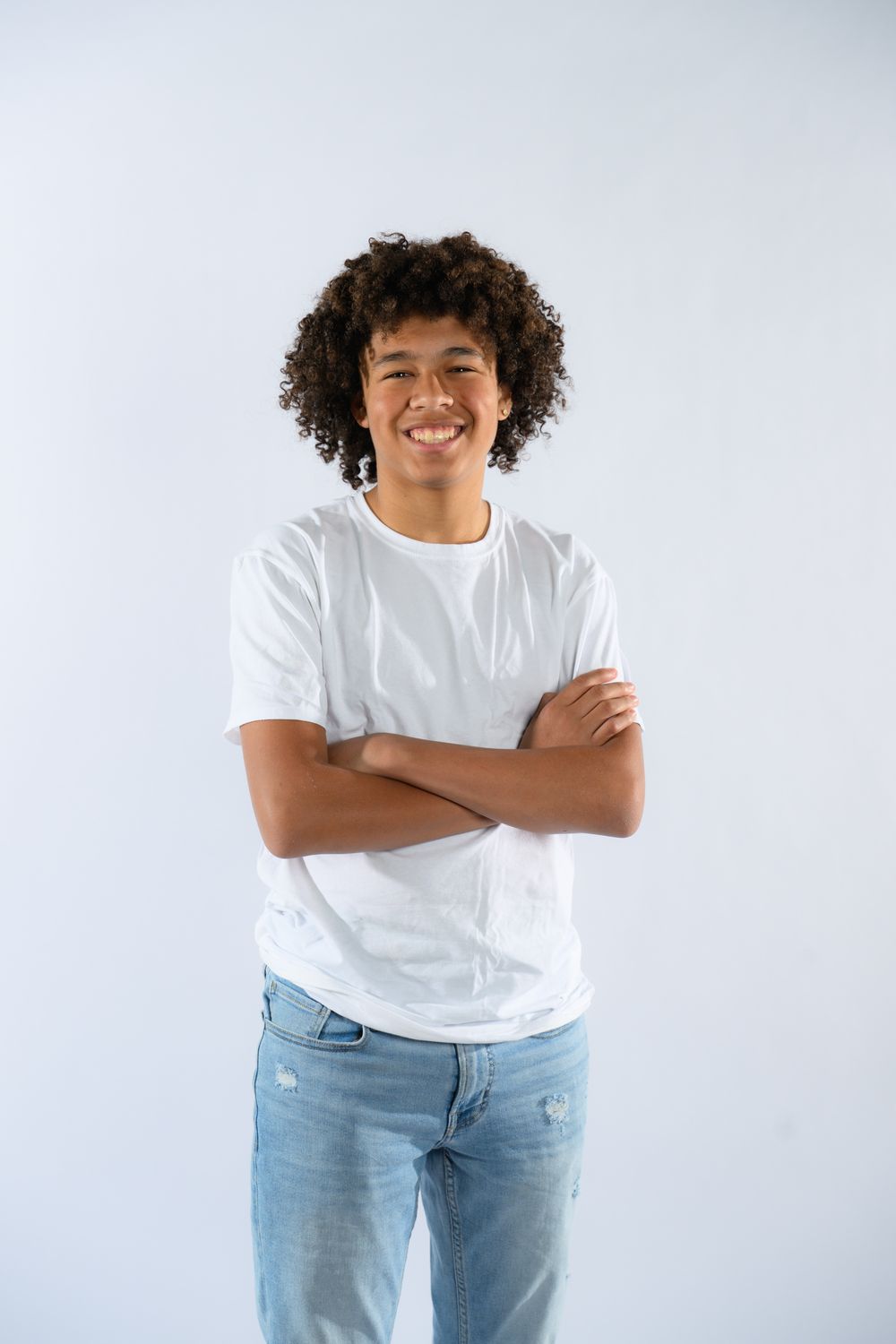 Smiling young man with curly hair wearing a white t-shirt and jeans, standing with arms crossed on a white background.