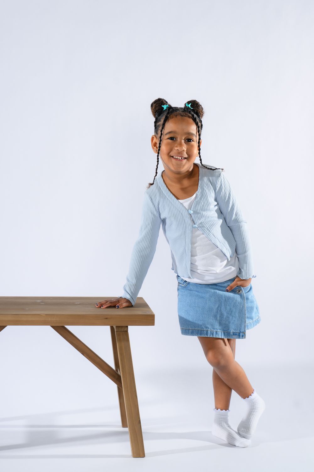 Smiling young girl in denim skirt and light blue cardigan standing next to a wooden table on white background.
