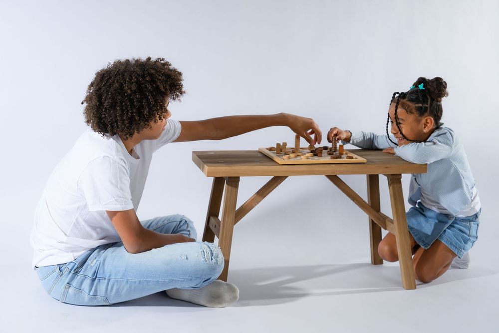 Two children playing chess at a wooden table, sitting on the floor against a white background.