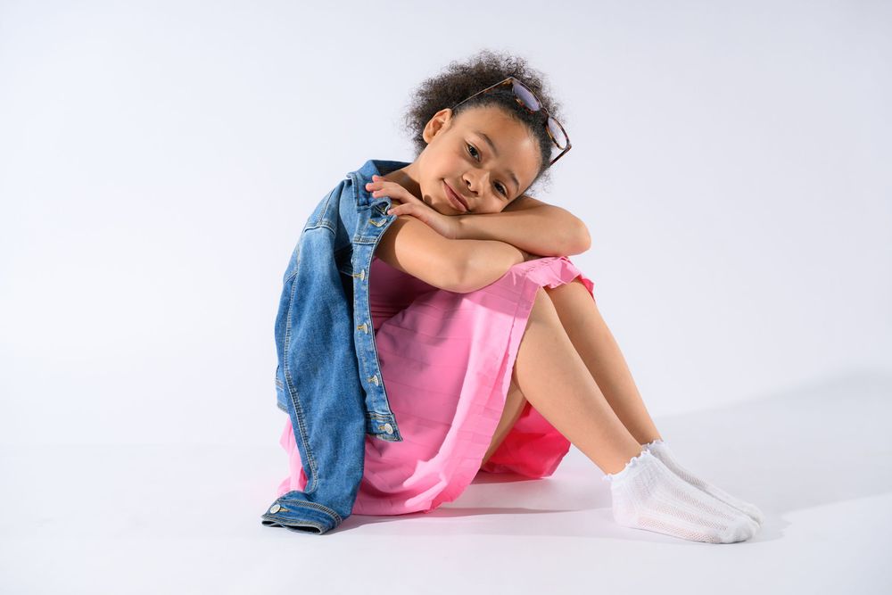 Young girl in pink dress and denim jacket sitting on white floor hugging her knees.