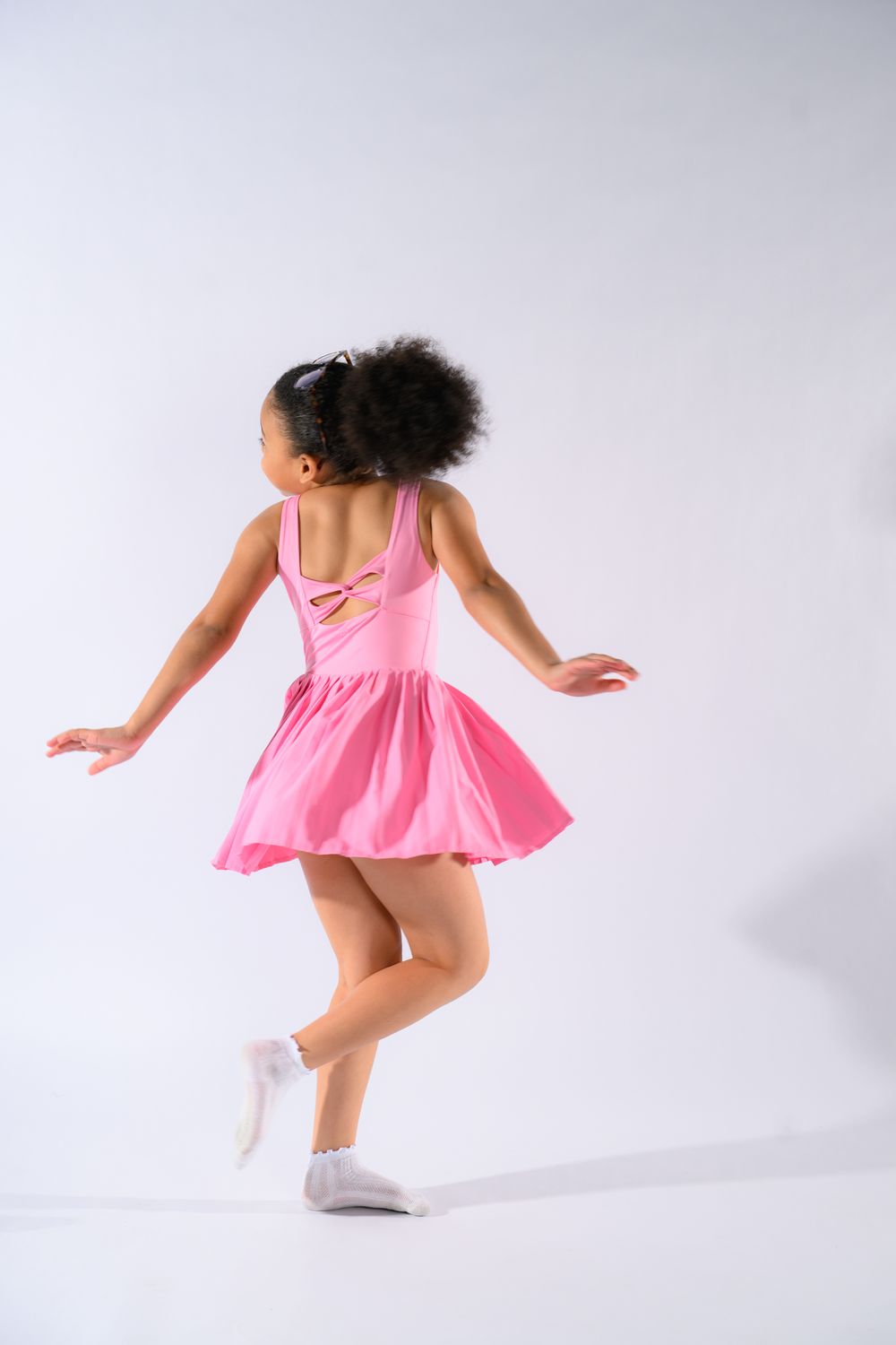 Young Black girl in pink ballet dress and white socks dancing gracefully against white background.