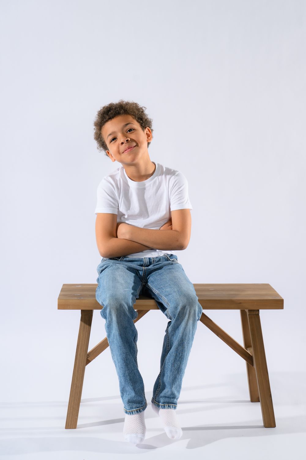 A smiling young boy in a white t-shirt and jeans sits on a wooden bench with arms crossed on white background.