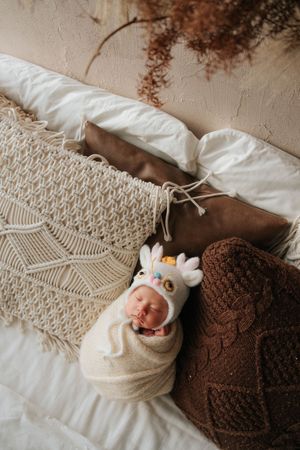 香港專業 newborn photographer 拍攝Newborn baby swaddled in cream wrap wearing a knit owl hat, sleeping peacefully among boho pillows and blankets.