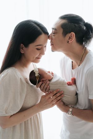 香港專業 newborn photographer 拍攝作品Asian couple in white clothing tenderly holding their swaddled newborn baby together.
