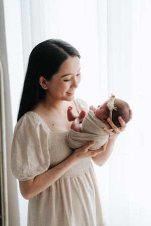 初生女兒與父母的溫暖互動畫面A young woman in a beige dress tenderly holds her swaddled newborn baby near a bright window.