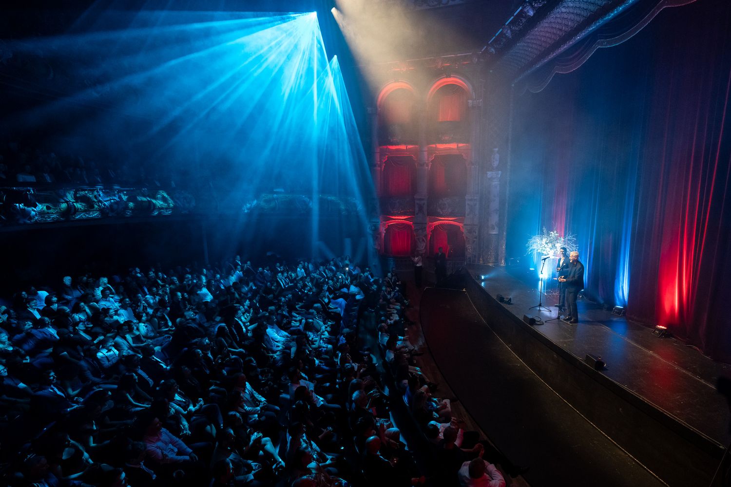 An interior venue photo inside the St James Theatre with Jamie Landau and James Cameron speaking on stage. The room is lit by blue & red.