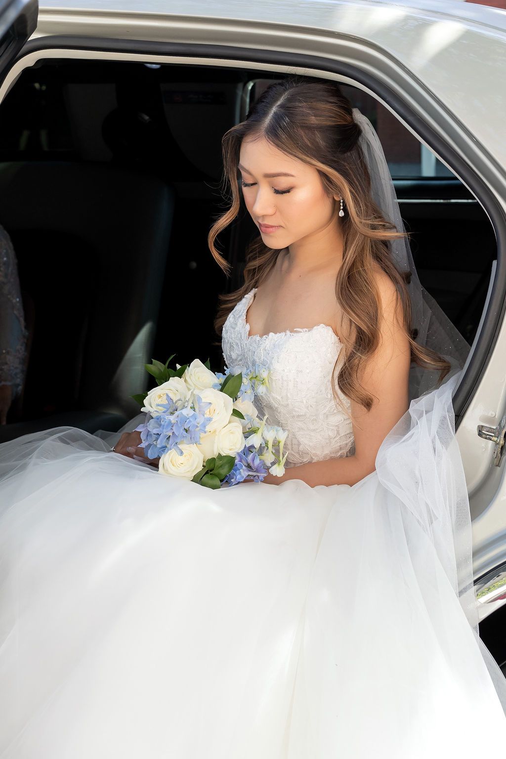Beautiful Asian bride in a white ballgown holding a blue and white bouquet inside a limousine.