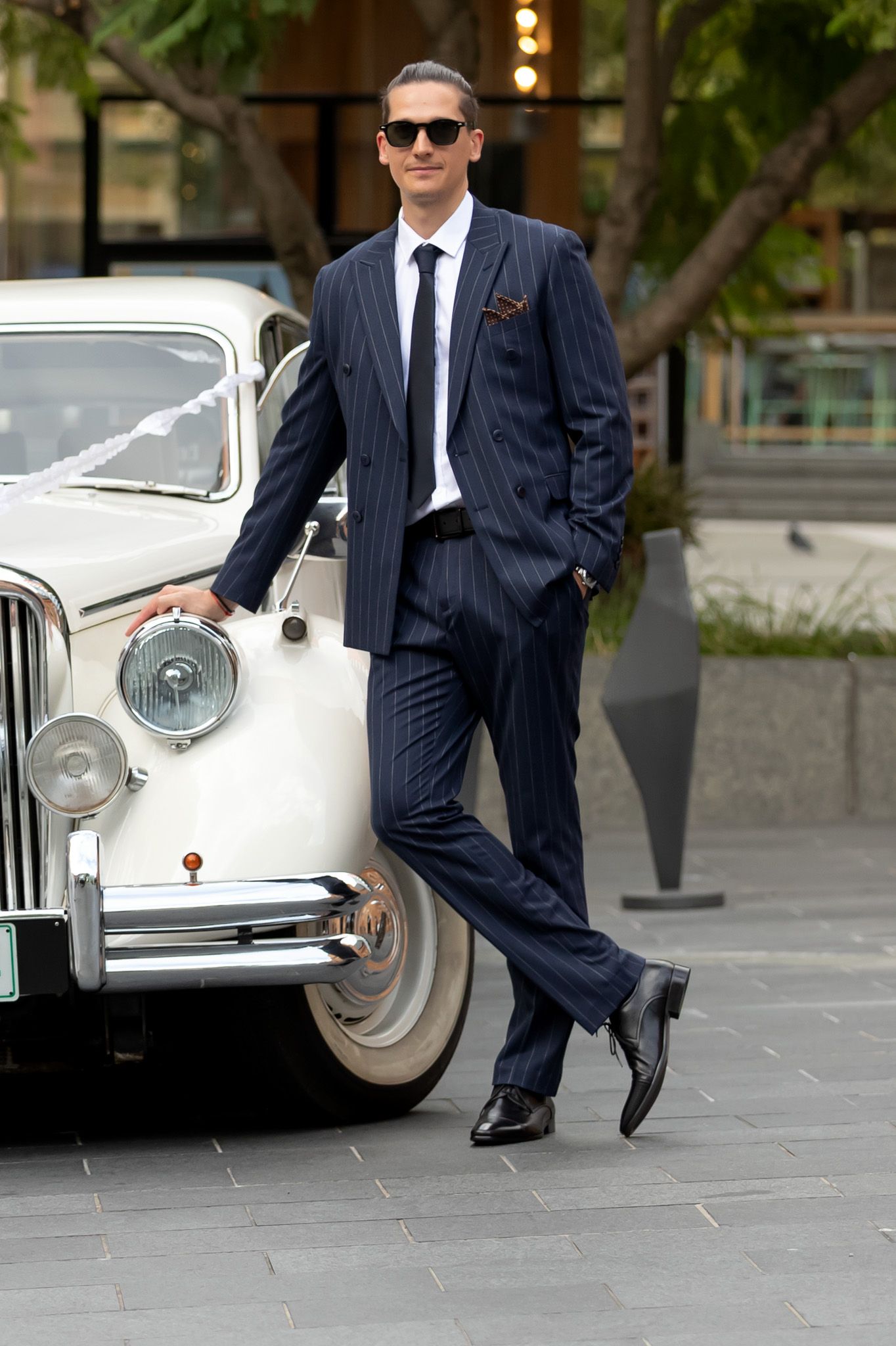 Stylish groom in a navy pinstripe suit leaning against a classic white vintage car wearing sunglasses.