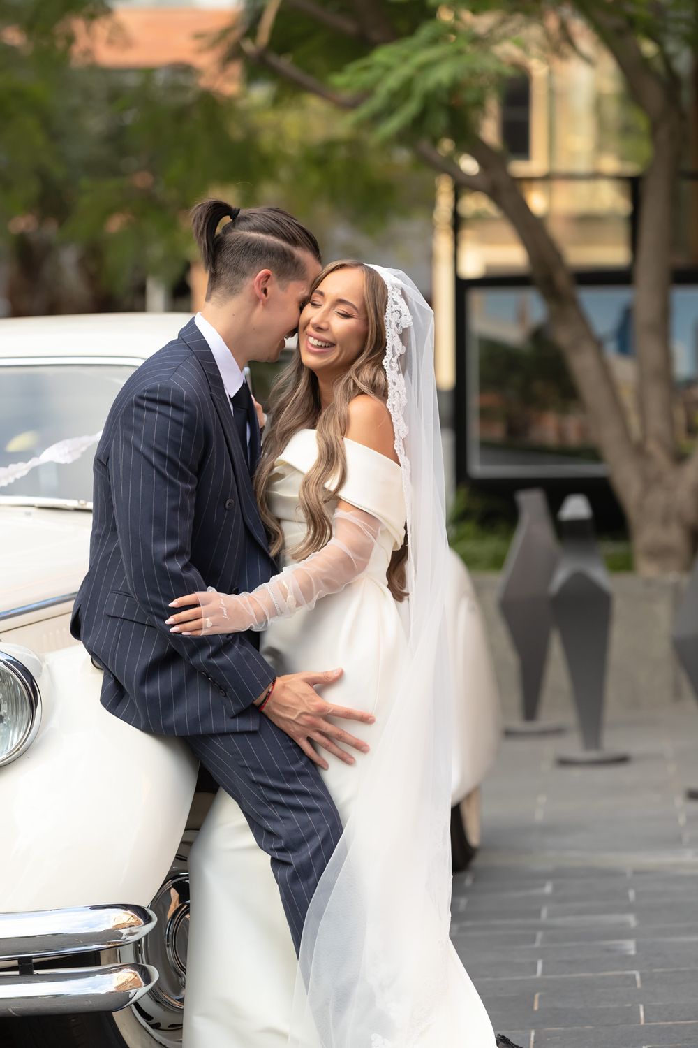 Bride and groom embracing and laughing together beside a vintage white car on their wedding day.