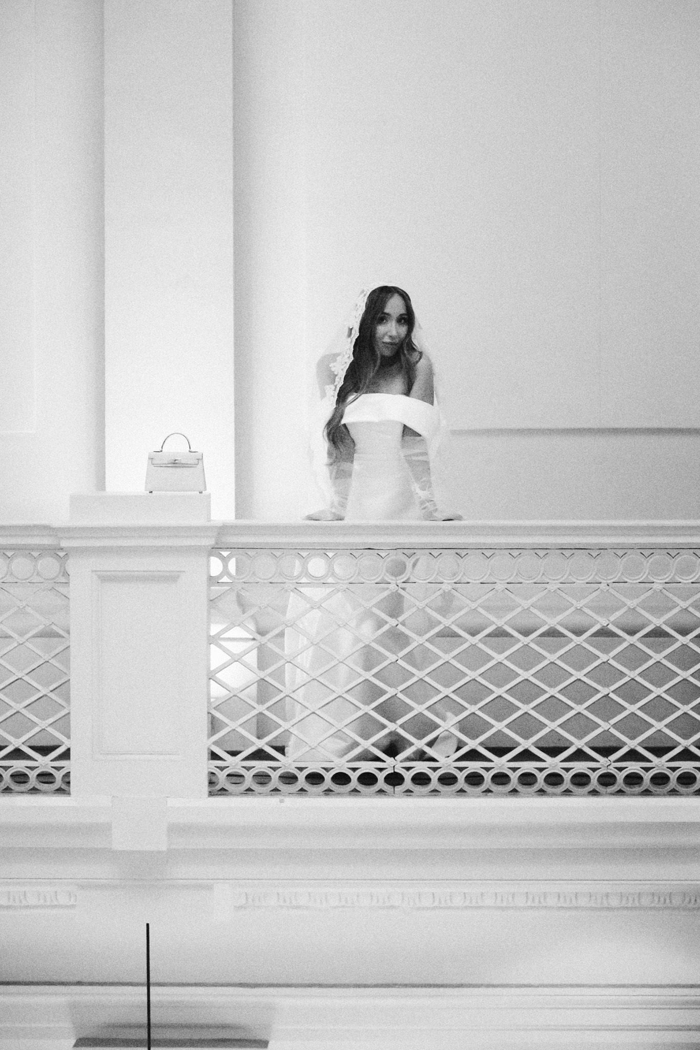 Black and white photo of a bride in a white gown standing on a balcony with ornate railings.