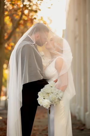 Bride and groom sharing a romantic kiss under a flowing white veil with autumn trees in the background.