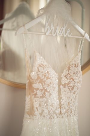 Intricate lace and beaded wedding dress hanging on a bride hanger in front of a mirror.