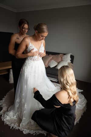 Two bridesmaids in black dresses helping bride into her lace wedding gown in a hotel room.