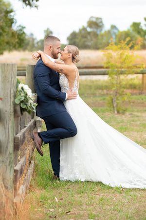 Bride and groom sharing an intimate moment leaning against a rustic wooden fence in a countryside setting.