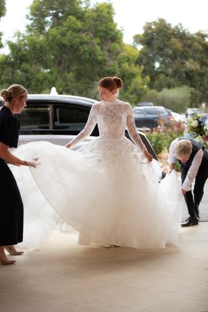 Bride in long-sleeve lace ball gown being assisted with her train outside a venue.