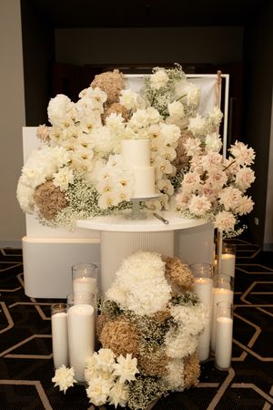 Elegant white wedding cake display surrounded by white and blush floral arrangements and candles.