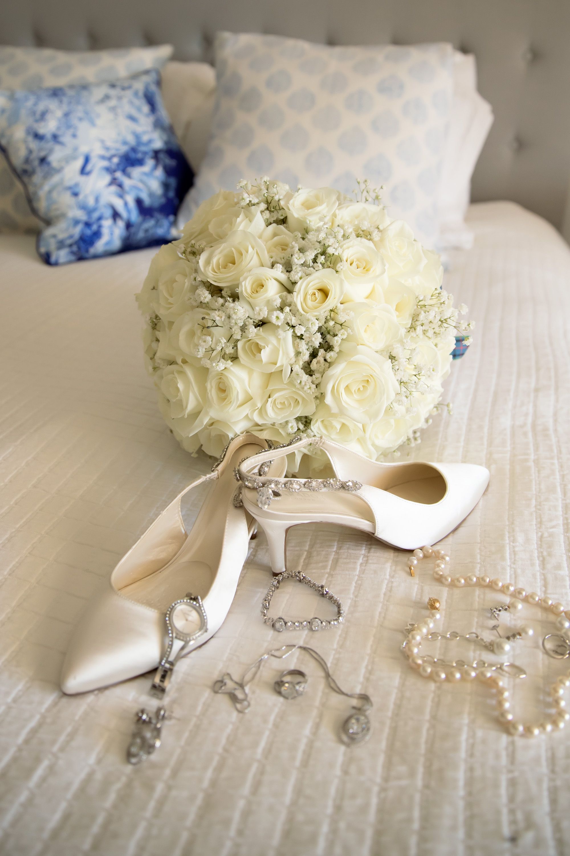 White rose bridal bouquet with pearl necklace, crystal jewelry and satin heels arranged on a bed.