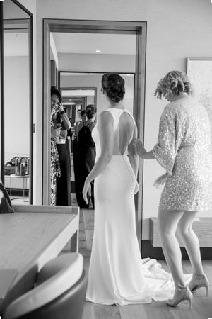 Black and white photo of bride in sleek backless gown being helped by bridesmaid in hotel room.