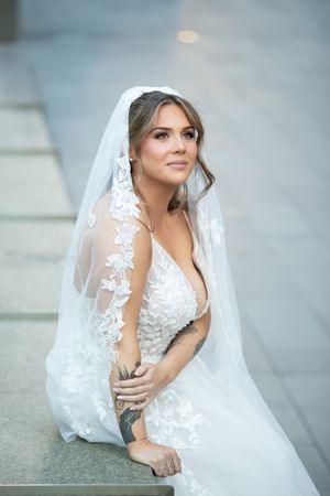 Elegant bride in deep V-neck lace wedding gown with floral veil sitting gracefully outdoors.