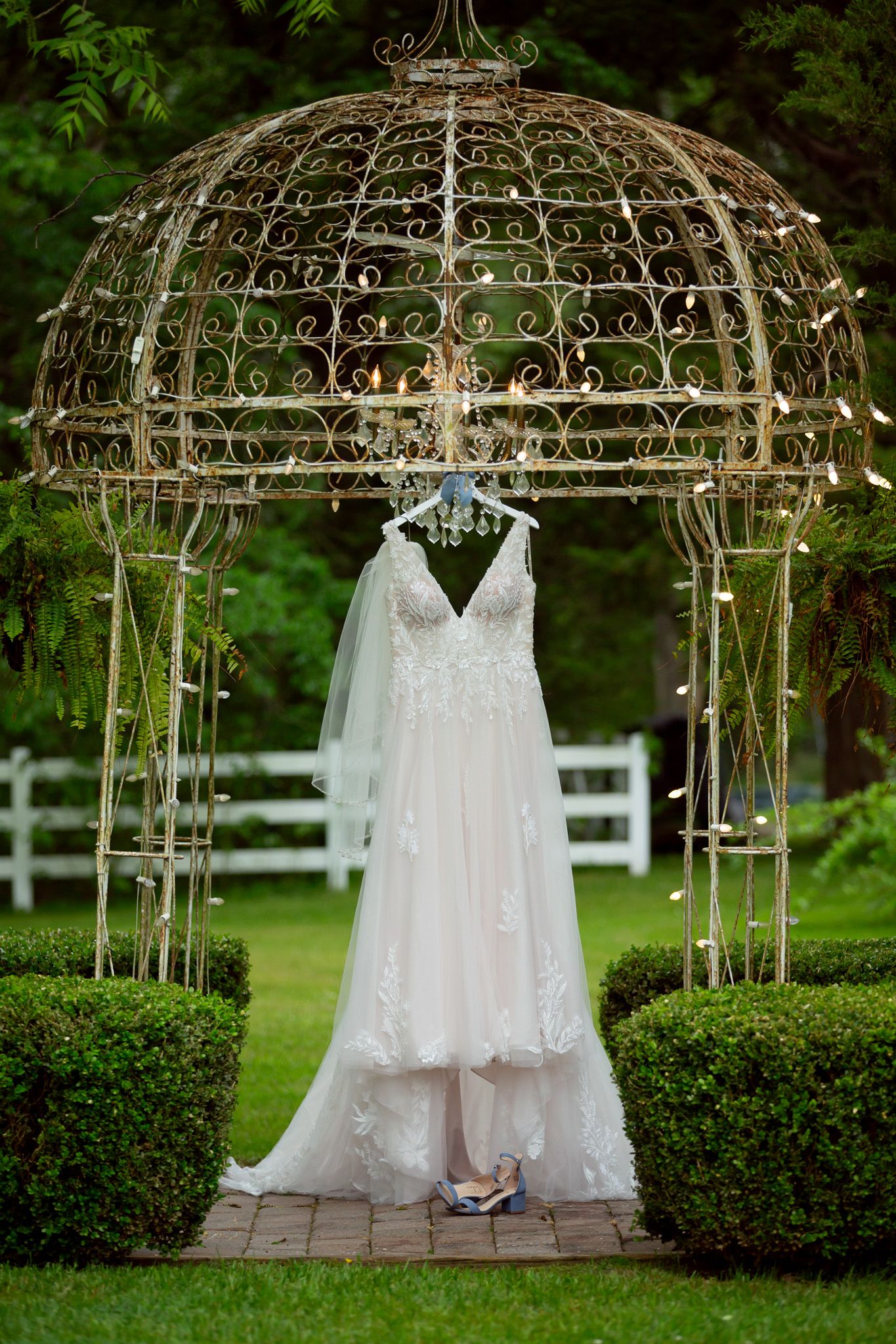 A lace wedding dress and veil hanging elegantly under a decorative iron gazebo in a lush garden setting.